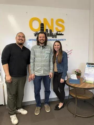 Three people smile for a photo under a sign saying ONS