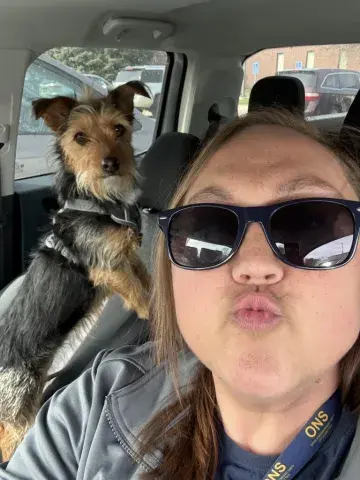 A woman in sunglasses takes a selfie with a small dog in a car.