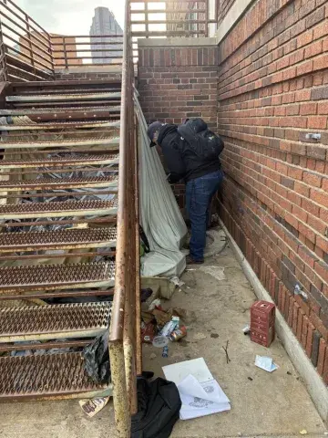 A person bends down to look inside a makeshift shelter constructed under an outdoor staircase.