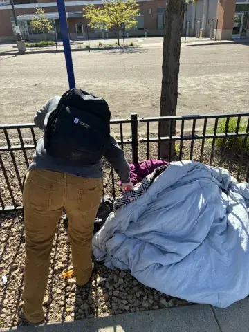 A person bends down to touch someone who in a sleeping bag on a city sidewalk.
