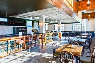 Sunlit indoor room with high ceilings. There are tables along the wall and bar stools along a counter, which also has a register and menu mounted above it.