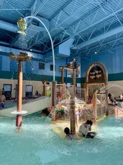 Young children play in a shallow indoor pool. There are several water features, including multiple fountains, and a play structure.