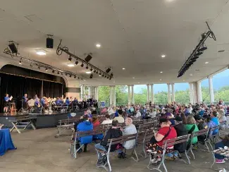 Large covered outdoor promenade looking out on the lake. A crowd of people sits on benches walking musicians perform on stage.