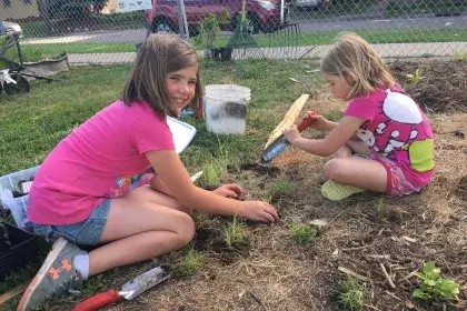 Two girls sit on the ground with trowels installing small plants