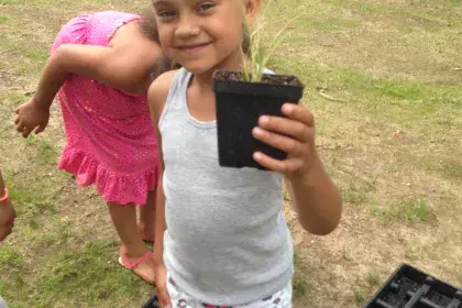 A child holds up a black plastic pot with a small plant inside