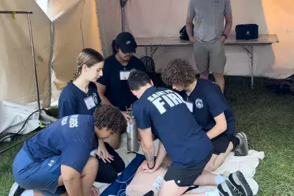Five teenagers crouch over a CPR dummy laid on a towel on the ground to practice CPR. The teens are wearing Saint Paul Fire Explorers shirts.