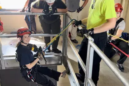 A smiling teenage girl wearing a helmet and harness descends from the second story of an atrium using a belay system while three adults look on. 