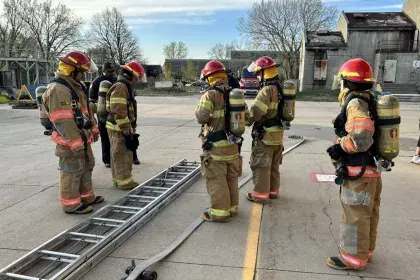 A group of teenagers in fire safety gear stand outside a training facility and prepare to put up a ladder.