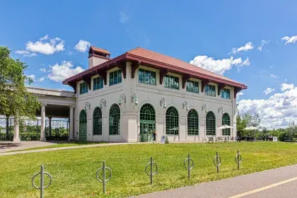 Front of the Como Lakeside Pavilion, with a large grassy lawn in front. There are multiple walking and bike trails passing by. A covered terrace is visible off the back of the building, with open access from the paths in front.