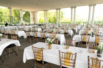Rows of tables with white tablecloths, flowers, and place settings on the covered, paved terrace of Como Lakeside Pavilion. The terrace is encircled by white columns and looks out onto Como Lake and surrounding trees.