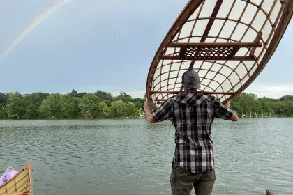 A man holds a lightweight canoe on his shoulders and approaches Como Lake. He stands on a paved brick area along the water's edge.