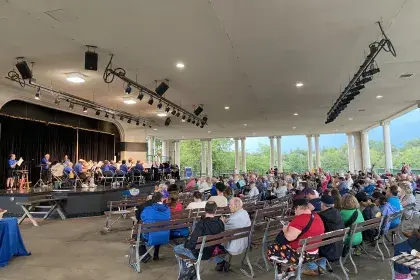 Crowds of people sit on benches on a large, paved outdoor terrace. They watch musicians perform on a stage which faces out toward Como Lake.