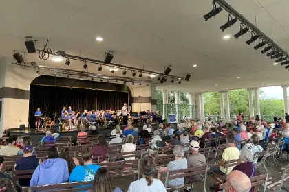 Crowds of people sit on benches on a large, paved outdoor terrace. They watch musicians perform on a stage which faces out toward Como Lake.