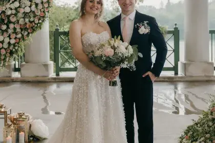 A couple poses for a wedding photo under a decorative arch on the paved, covered terrace of Como Lakeside Pavilion. White columns and the lake are in the background.