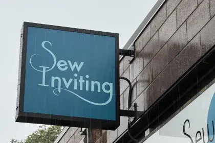 Blue blade sign for Sew Inviting mounted on a brick storefront on a rainy day.