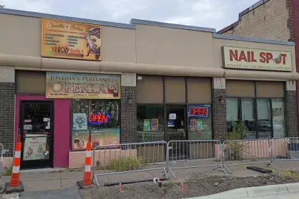 Row of small businesses on a construction-affected block, including Claudia's Salón, Joyería y Perfumería Perla and Nail Spot, with construction barricades and torn-up sidewalks in front.